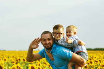Fototapeta premium Cheerful father with his sons on vacation in the field with sunflowers . The parent plays with his son . Father's day