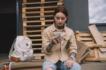 stylish tattooed woman in earphones listening music with smartphone near skateboard and backpack