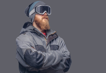 Portrait of a redhead snowboarder with a full beard in a winter hat and protective glasses dressed in a snowboarding coat posing at a studio, looking away. Isolated on gray background.