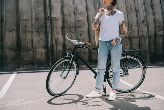 cropped image of stylish tattooed woman holding jacket over shoulder near bicycle at street