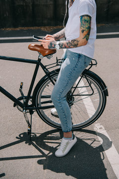 cropped image of tattooed woman in earphones listening music with smartphone while sitting on bicycle