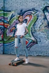 young tattooed woman standing on skateboard near wall with colorful graffiti