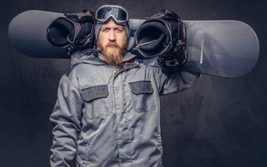 Brutal redhead snowboarder with a full beard in a winter hat and protective glasses dressed in a snowboarding coat posing with snowboard at a studio. Isolated on gray background.