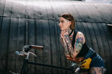 Fototapeta na wymiar smiling stylish tattooed girl standing with bicycle at street