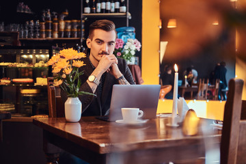 Portrait of a pensive handsome businessman with stylish beard and hair dressed in a black suit sitting with hand on chin at a cafe with an open laptop, lookong at the camera.