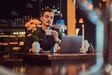 Handsome freelancer man with stylish beard and hair dressed in a black suit sitting at a cafe with an open laptop and holds a cup of coffee, looking at the camera.