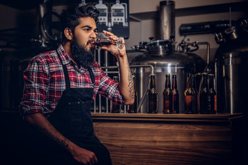 Bearded tattooed Indian hipster male in a shirt and apron working in a brewery factory, sitting behind a counter, drinks craft beer for quality control.