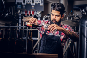 Stylish full bearded Indian man in a fleece shirt and apron pours beer in a glass for quality control, standing behind the counter in a brewery.