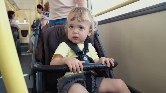 Side view of adorable little kid in light summer clothes sitting in baby stroller in bus and looking at camera