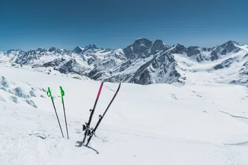 Fotobehang A pair of skis and ski poles stick out in the snow on the mountain slope of the Caucasus against the backdrop of the Caucasian mountain range and the blue sky on a sunny day © yanik88