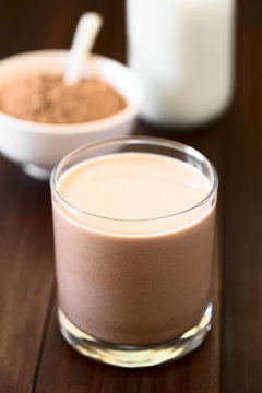Chocolate milk drink in glass, chocolate or cocoa powder and milk in the back, photographed with natural light (Selective Focus, Focus on the front of the glass rim)
