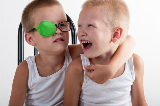 Two happy children play together in room. Boys brothers on white background. Сhild in glasses with green occluder. Ortopad Boys Eye Patches Nozzle for glasses to treat strabismus (lazy eye)