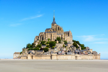 General view of the Mont Saint-Michel tidal island, located in France on the limit between Normandy and Brittany, with the exposed sand of the bay at low tide in the foreground under a blue sky.