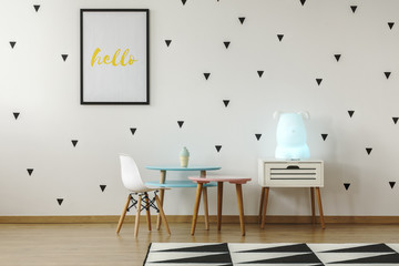 White chair next to pastel tables in bright child's room interior with poster and carpet. Real photo