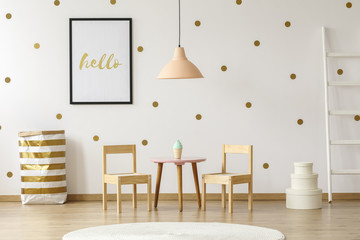 Pastel lamp above table and wooden chairs in white and gold kid's room interior with poster. Real photo © Photographee.eu