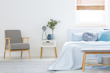 Plant on white cabinet between patterned armchair and blue bed in bedroom interior. Real photo