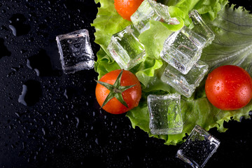 Fototapety  Banda czerwonych pomidorów cherry, zielonej sałaty i kostek lodu na czarnym mokrym stole. Selektywne skupienie