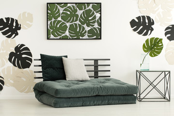Poster of leaves above futon with cushions in modern bedroom interior with table. Real photo