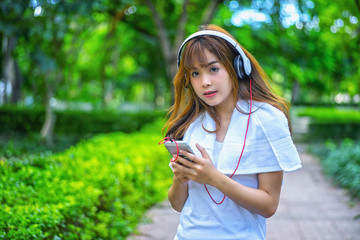 Young happy smiling asian woman listening music with headphones from her smart phone at the park during a sunny day wearing white clothes