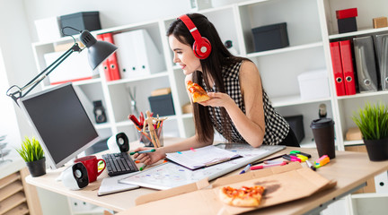 A young girl is standing near a table, holding a green marker and a piece of pizza in her hand. Before the girl On the table is a magnetic board. On the head of the girl wearing headphones.