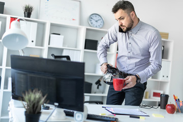 A young man in the office is standing near the table, holding the phone with his shoulder, looking at the monitor and pouring coffee into the cup.