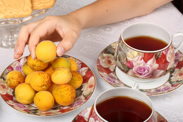 A teenager takes a wild apricots from a plate. Teacups with hot tea and with floral pattern, teapot, biscuits and plate with wild apricots