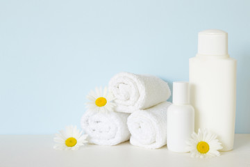 Fototapeta na wymiar White bottles and towels on white shelf in bathroom. Beautiful camomiles or daisies. Fresh flowers. Care about clean and soft face, hands, legs and body skin. Empty place for text on pastel blue wall.