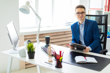 A young man sits at a table in the office and holds documents and a pen in his hands.