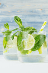 Fresh cold summer beverage gin and tonic with lime, leaf mint, straw, ice cubes, soda on light blue wooden background, closeup.