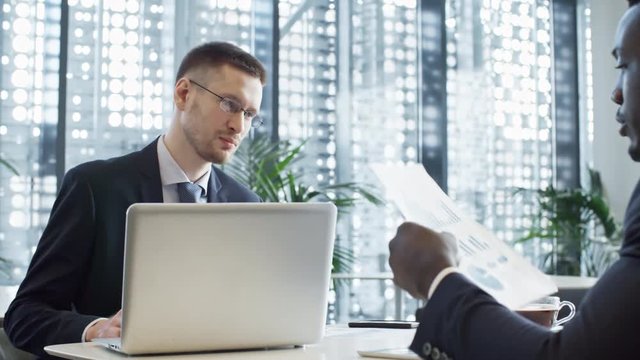 Young Caucasian businessman reading financial documents while working with African American colleague in large office