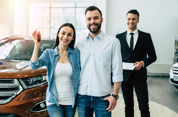 We bought a car. Young and smiling man and woman show the keys to a new jeep looking at the camera on the background of the dealership