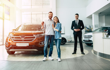 Happy smiling couple with keys in their hands from a new car looks at the camera in the dealership.