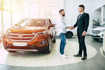 The manager in the dealership shakes hands with a young man in a light shirt against a new car.