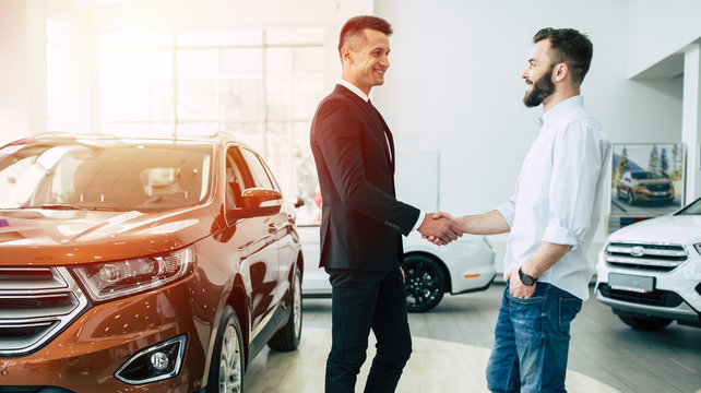 The manager in the dealership shakes hands with a young man in a light shirt against a new car.