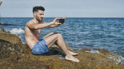 Young handsome man at the seaside using smartphone to take photos and film footage of the sea or ocean