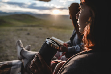 two young girls sitting in the van, reading a book and drinking from a thermos. beautiful sunset in mountain valley on the background.
