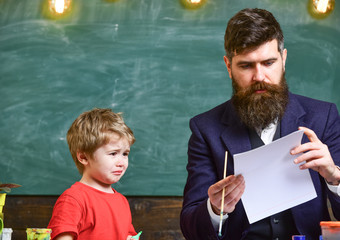 Teacher and kid in classroom with chalkboard on background. Family on busy faces creating, drawing. Father and son painting together in study room.