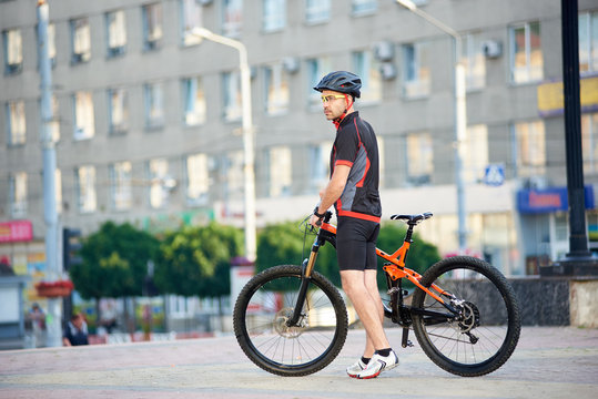 Young man in relaxing after bike ride standing near bicycle in front of city building. Cyclist looking far and posing for sport garment ad campaign. Concept of healthy lifestyle