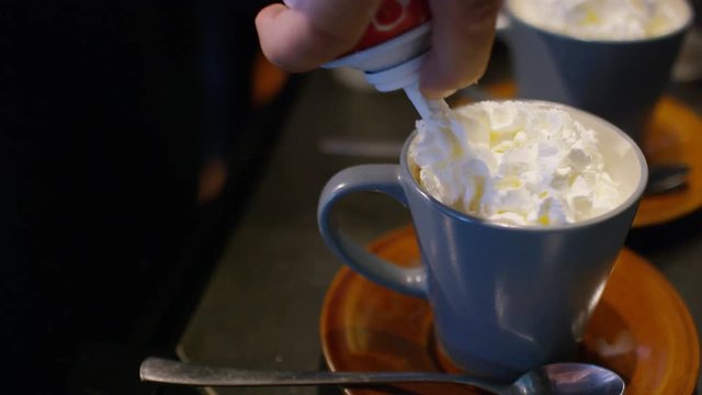 Whipped cream being sprayed onto mugs of hot chocolate followed by marshmallows