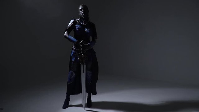 Cosplay of woman dark warrior that stands on herheels and holds a magic sword on her shoulder
