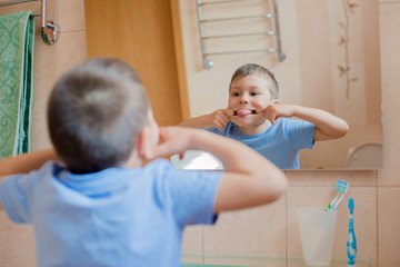 The child grimaces before the mirror in the bathroom. A little boy shows his tongue and has fun.