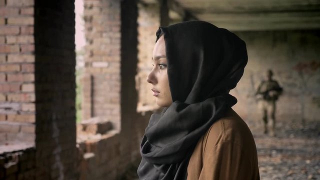 Young sad woman in hijab standing, soldier with ammunition coming towards woman, abandoned building background, terrorism