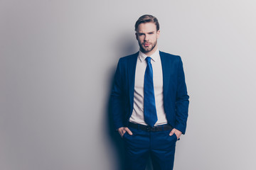 Fototapeta premium Portrait of stunning, attractive, sexy, brutal man in blue suit with tie with stubble, holding two hands in pockets of pants, looking ta camera, isolated on grey background