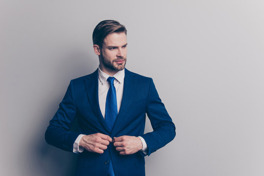 Portrait with copy space of stunning, fashionable, cool, virile, rich man with stubble in blue suit fasten button on jacket with two arms, looking to the side, isolated on grey background