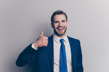 Portrait of attractive, laughing, cheerful, positive man in blue suit with modern hairstyle, stubble, showing thumb up sign with arm, looking at camera, isolated on grey background