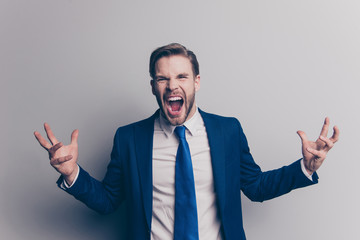 Portrait of violent, stylish, attractive, angry man in blue suit, tie with bristle, hairstyle, gesturing with twoarms, yelling with wide open mouth, looking at camera, isolated on grey background