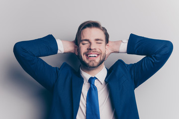 Portrait of glad, cheerful, positive man with close eyes in blue suit with tie, holding hands behind the head, enjoying, dreaming about vacation, weekend, isolated on grey background