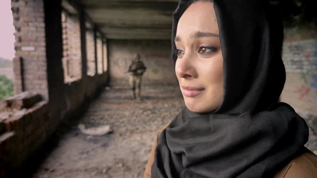 Young woman in hijab standing in abandoned building, soldier walking in background
