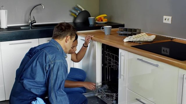 Tilt down of black woman talking and showing broken dishwasher machine to male plumber in overalls