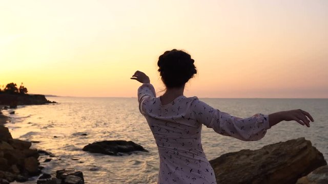 Adorable Caucasian Brunette girl in Light Dress Dancing at the Cliff above the Sea at Sunrise time